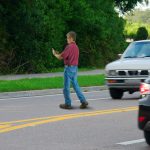 A man who is completely distracted by his cell phone is unaware that he is jaywalking by walking out into heavy traffic on a busy highway road.